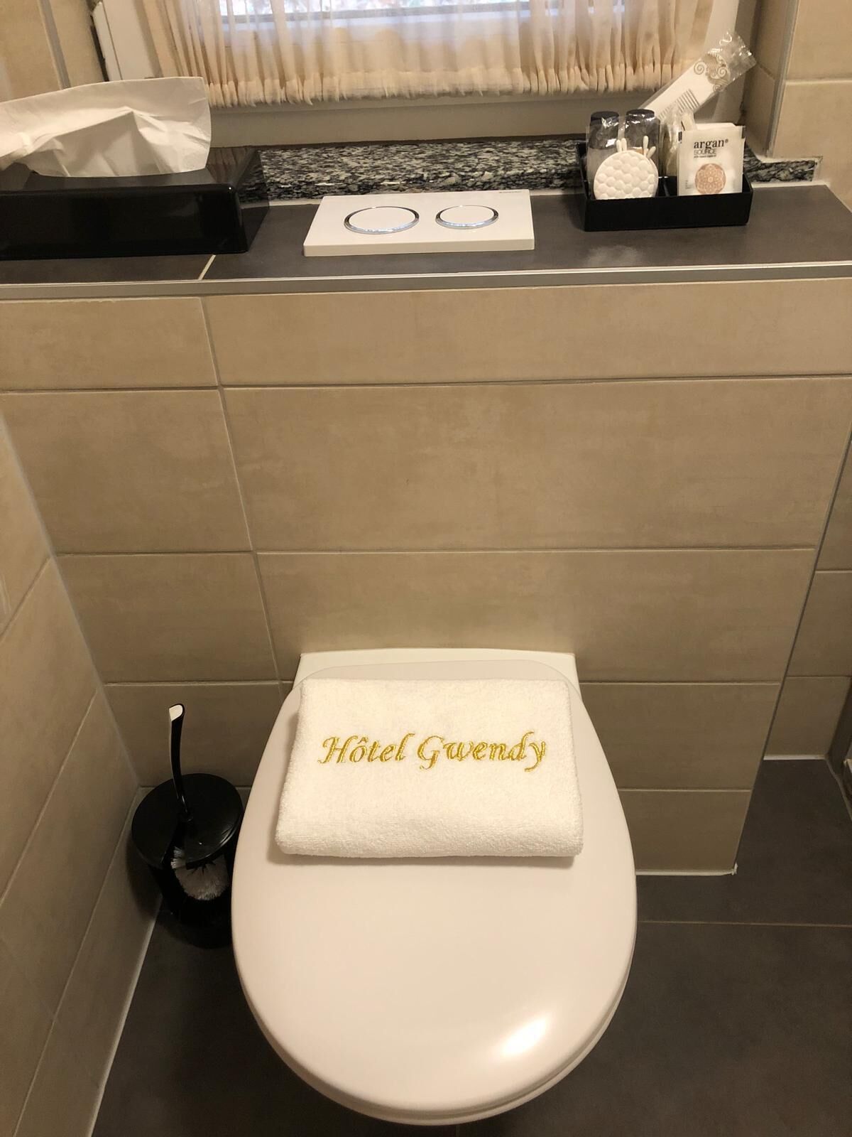 A bathroom with a white toilet, a towel with "Hotel Gwendy" embroidered, a black trash bin, and toiletries on a shelf.