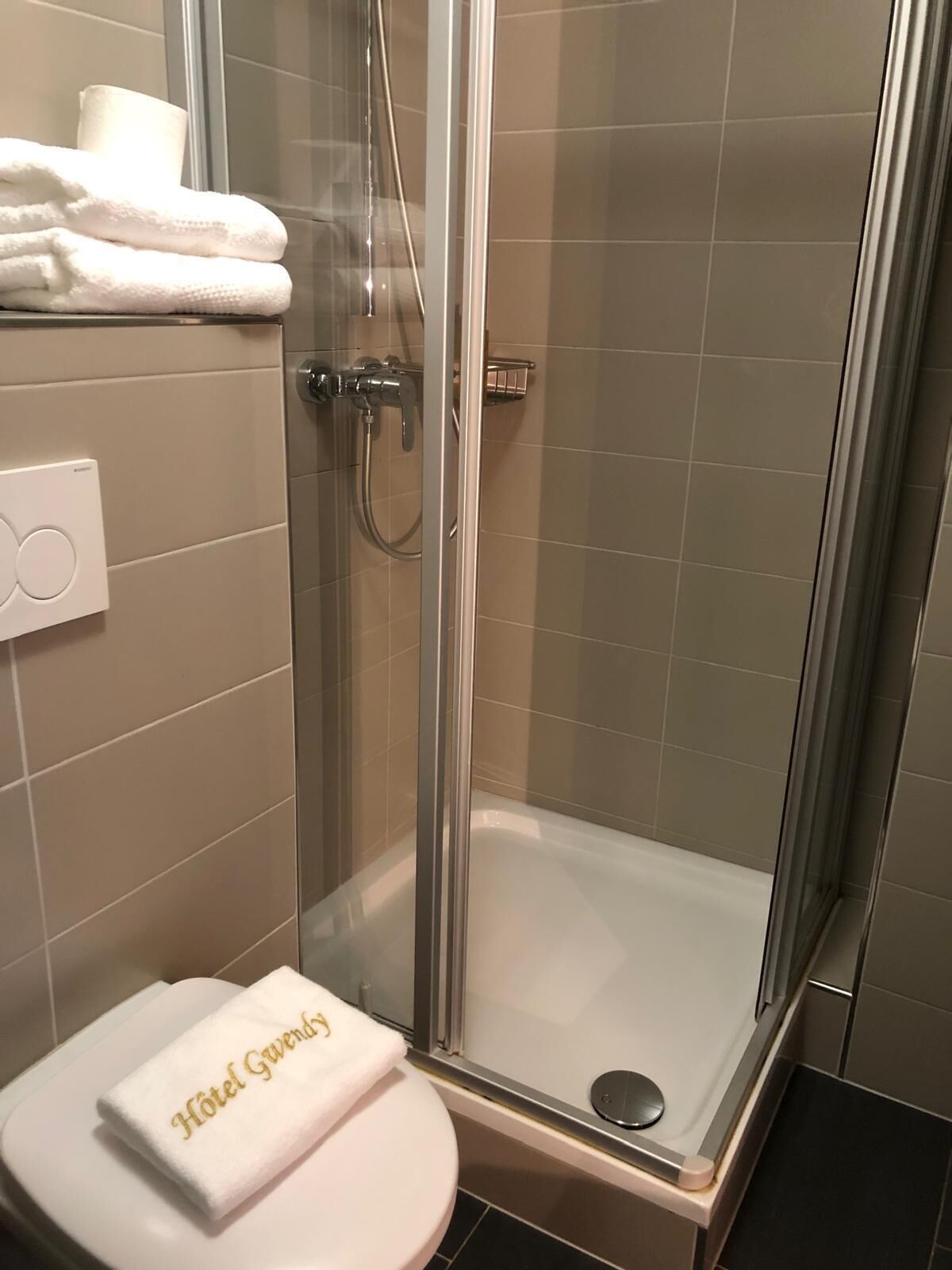 Une douche fermée par des parois de verre, avec des toilettes blanches et une serviette sur laquelle est brodé le nom « Hôtel Gründ ».