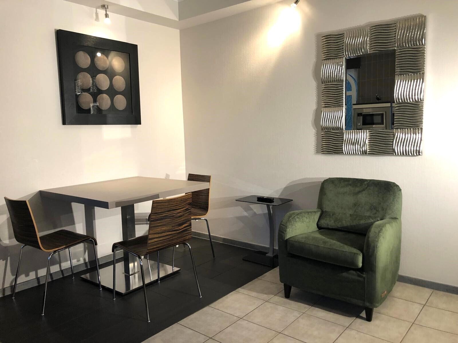 A modern living room with a dining table, two chairs, a green armchair, a small side table, and a decorative wall piece.