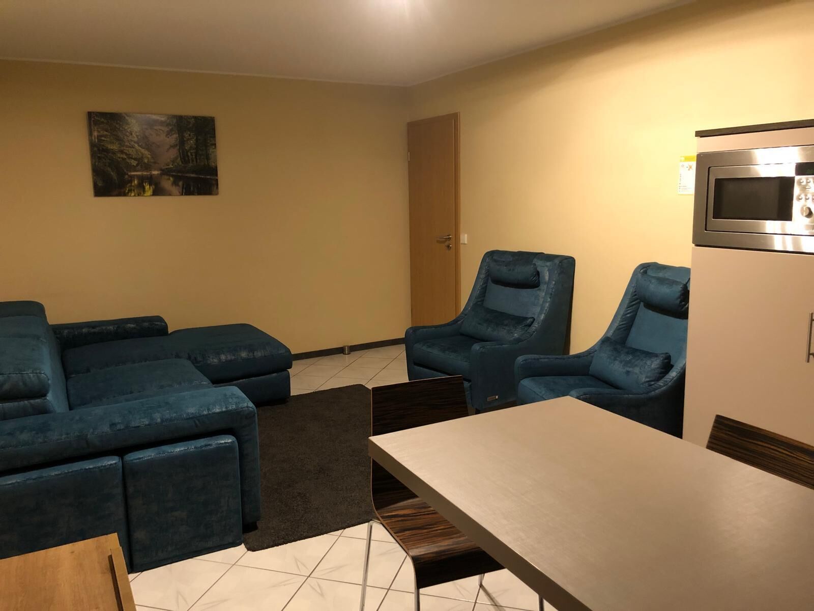 A living room with a sectional sofa, two armchairs, a coffee table, and a dining table. There is a painting on the wall and a microwave on the wall.
