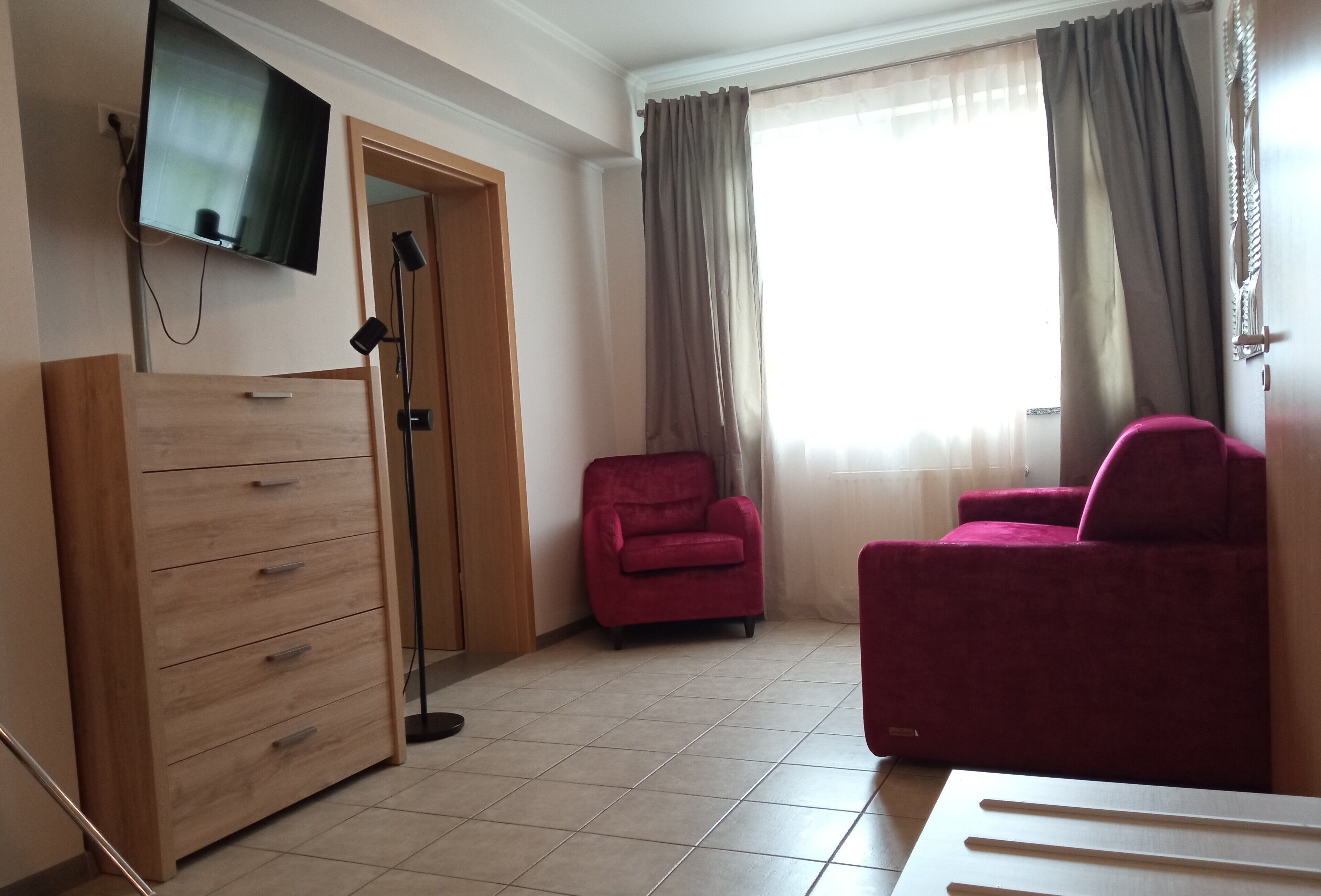 Een kamer met een houten dressoir, een rode fauteuil, een bank, een aan de muur bevestigde televisie en een raam met gordijnen.