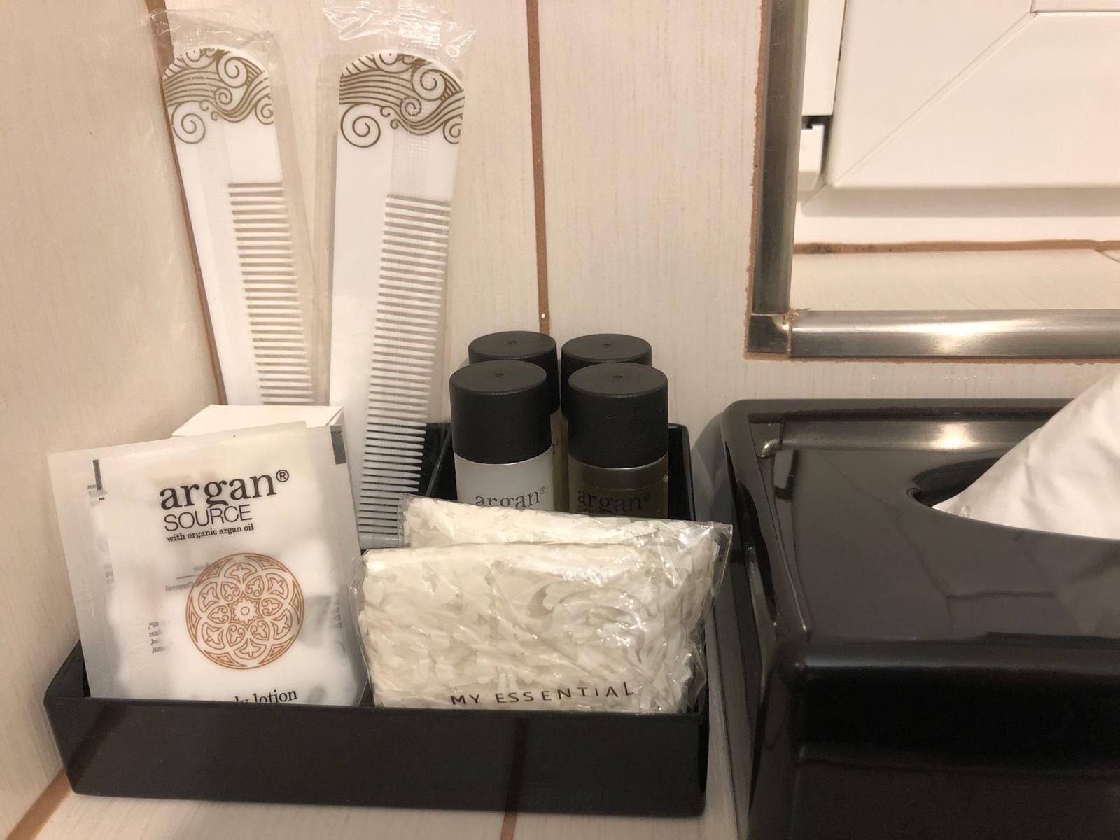 Servicios de la habitación de hotel, incluyendo un peine blanco, loción Argan Source y una bandeja negra con una caja de pañuelos.
