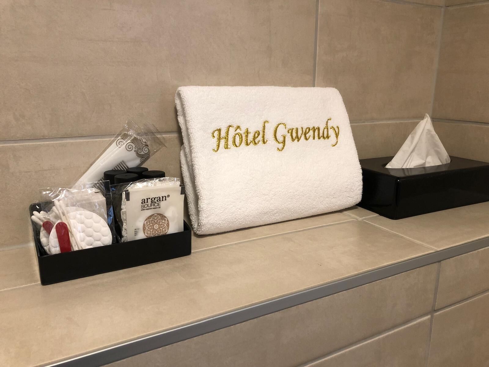 A hotel bathroom with a towel rack holding a white towel with "Hotel Gwen" embroidered in gold, a black tissue box, and a tray with toiletries.