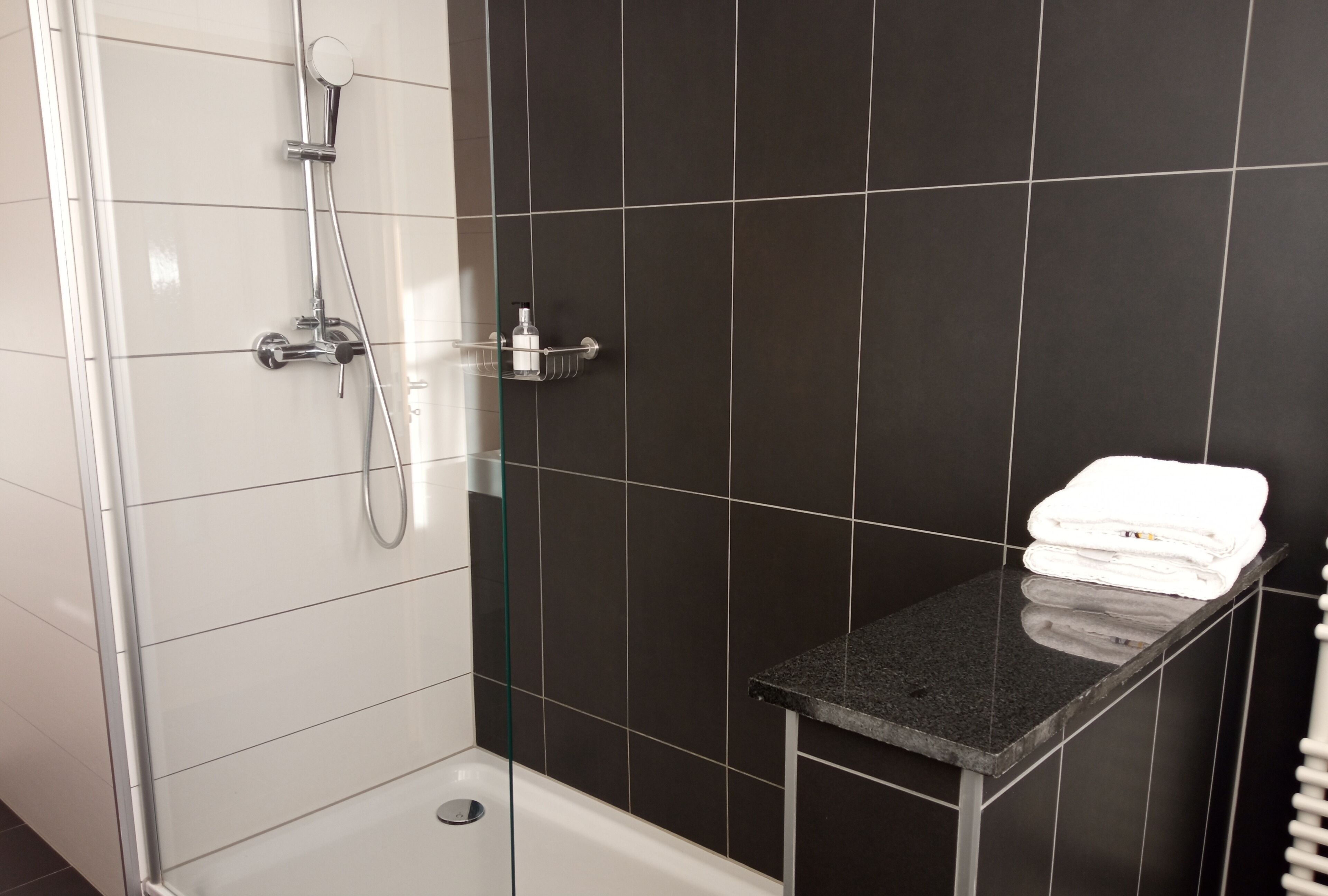 A modern bathroom with a glass-enclosed shower, a black tiled wall, and a small black countertop with a folded white towel.