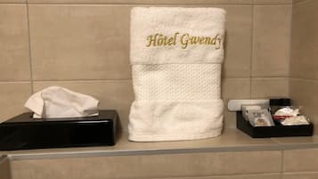 Un baño con un toallero blanco que sostiene una toalla bordada en dorado con la leyenda "Hotel Gwendy", una caja de pañuelos negros y una pequeña bandeja con artículos de aseo.