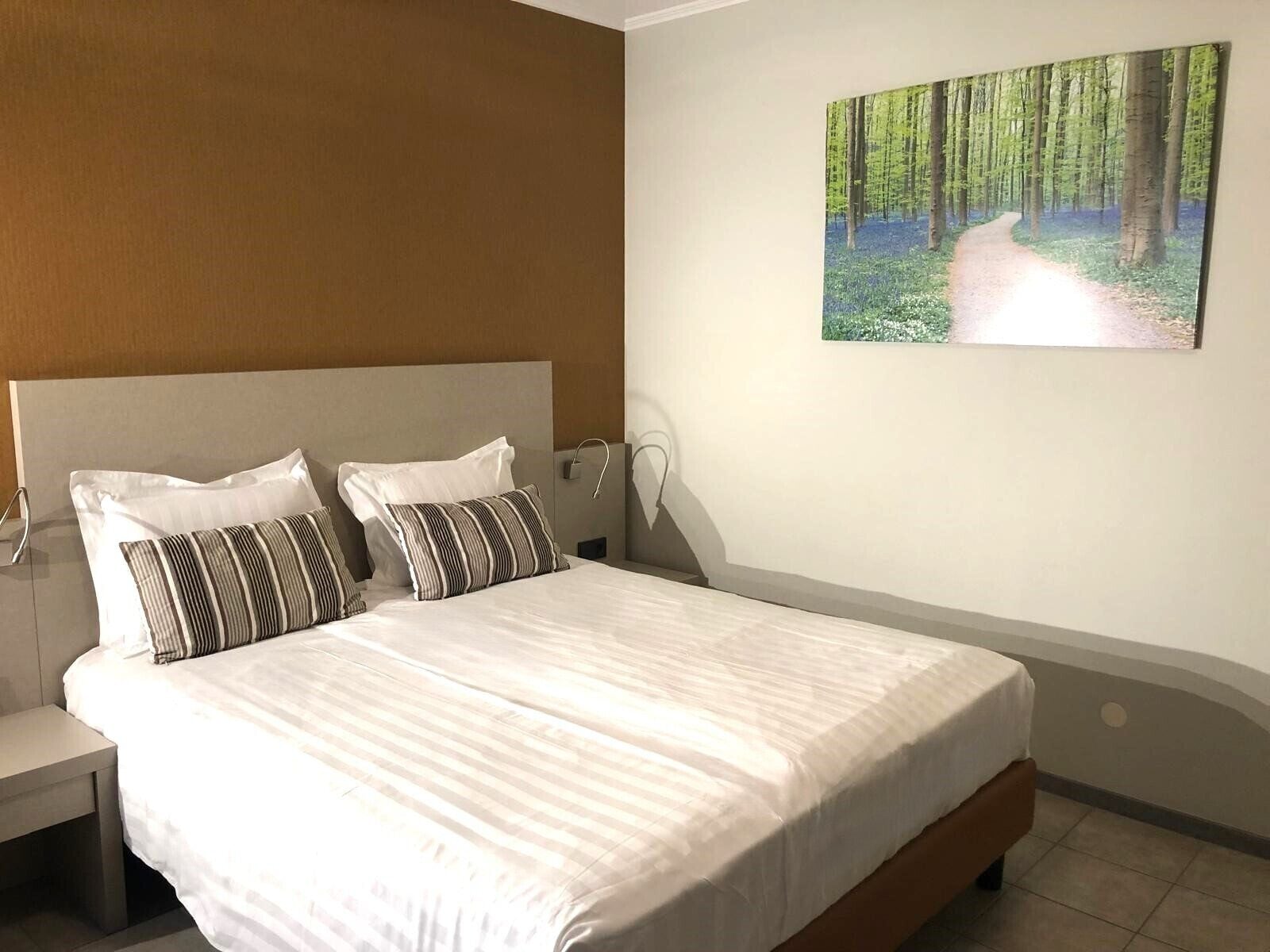 Un dormitorio con una cama grande, dos almohadas, una mesita de noche y un cuadro de un sendero forestal en la pared.
