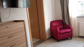 Une pièce comprenant une commode en bois, une chaise rouge, une porte en bois et un téléviseur à écran plat fixé au mur.