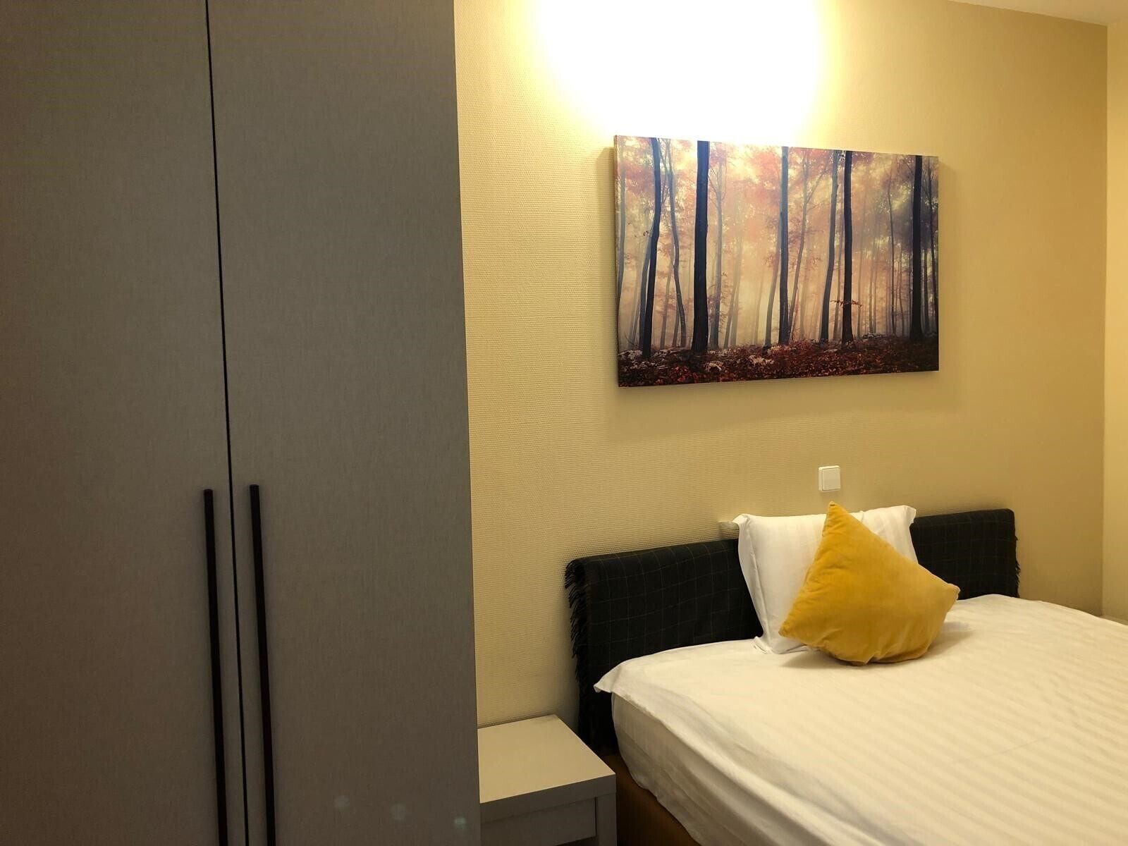 A bedroom with a bed, a nightstand, a wardrobe, and a painting of a forest on the wall.