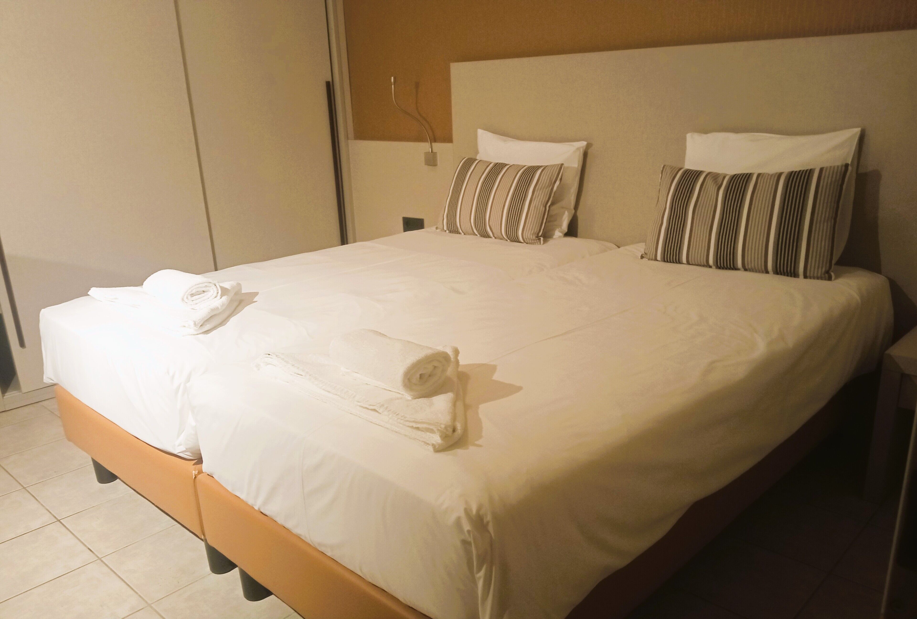 A neatly made bed with white linens and striped pillows, a rolled towel, and a wooden headboard.