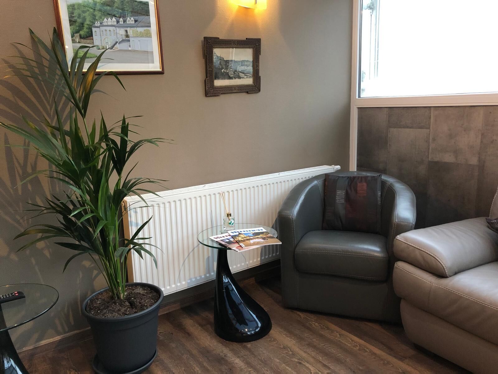 A cozy living room with a leather armchair, a sofa, a potted plant, a glass coffee table with magazines, and framed pictures on the wall.