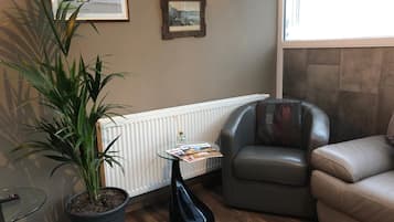 A cozy living room with a leather armchair, a sofa, a potted plant, a glass coffee table with magazines, and framed pictures on the wall.