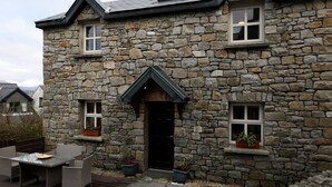 Ein Steinhaus mit einer schwarzen Tür, zwei Fenstern mit Blumenkästen und einer kleinen Terrasse mit Tisch und Stühlen.