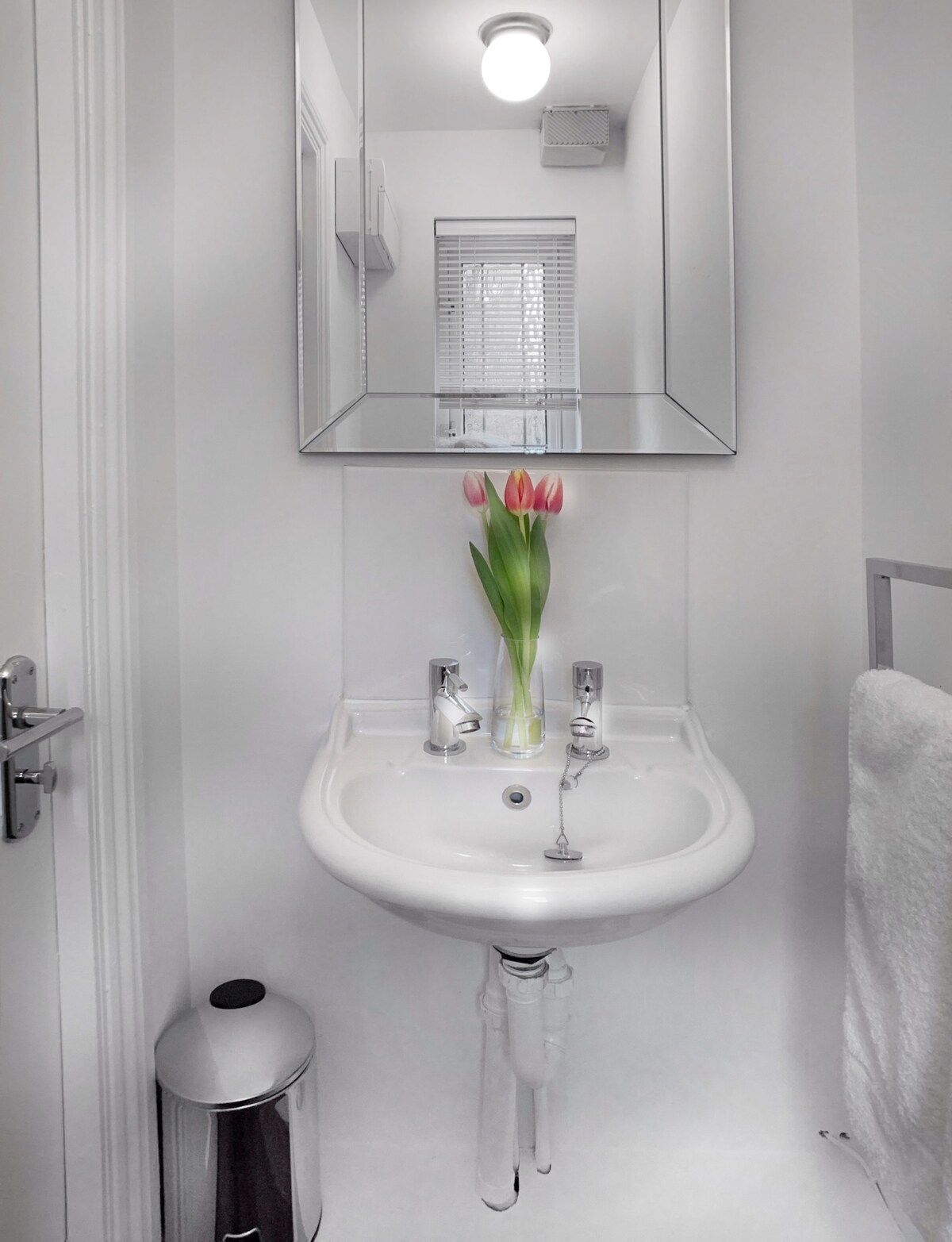 Ein Badezimmer mit einem weißen Waschbecken, zwei Wasserhähnen, einem Spiegel über dem Waschbecken und einem Fenster mit Jalousien.