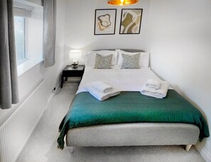 A neatly made bed with a green blanket and white pillows, a bedside table with a lamp, and abstract art on the wall.