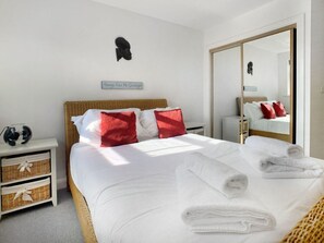 A neatly made bed with white linens and red pillows, a bedside table with a lamp, and a mirror reflecting the room. - Windsor - 1 Bedroom Flat - With Parking (Windsor)