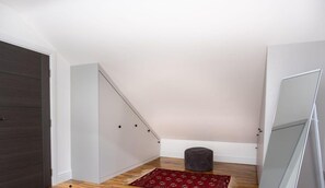 A room with a dark wooden door, a white wardrobe, a red patterned rug, a grey ottoman, and a large mirror on the wall.