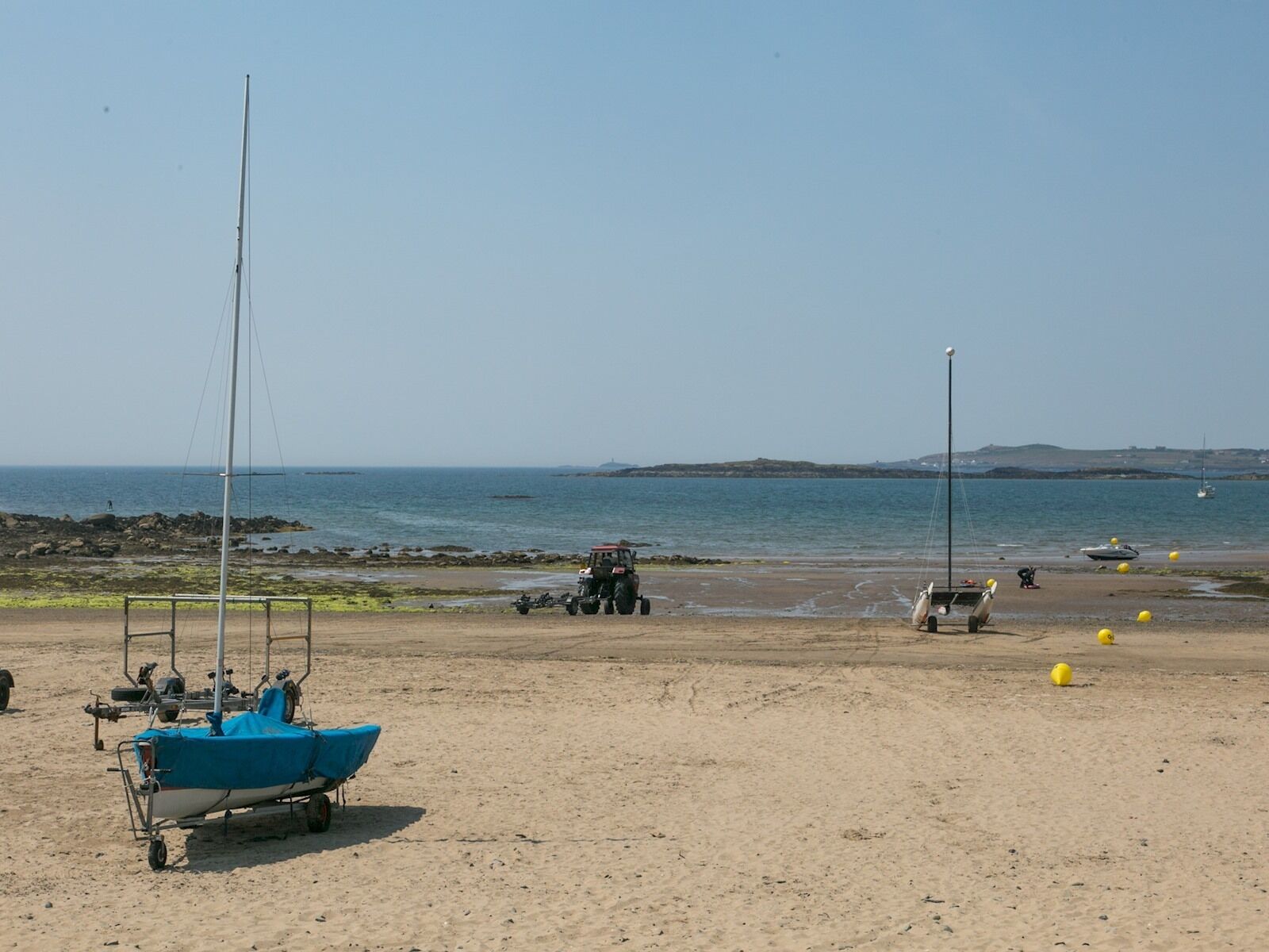 A sandy beach with a small boat on a trailer, a tractor, and several yellow buoys.