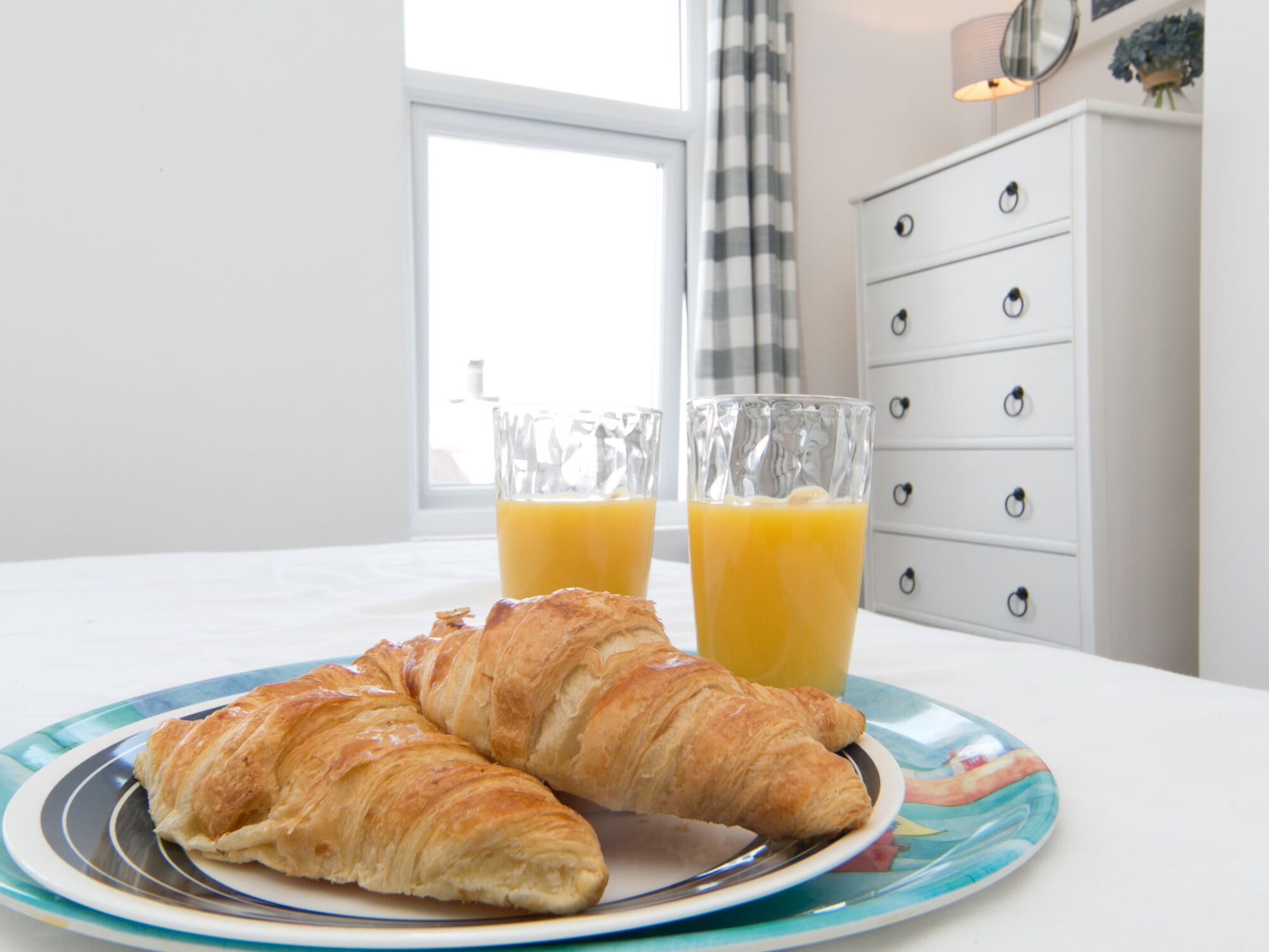 A breakfast setting with croissants and orange juice on a plate, a white dresser, and a window with curtains.