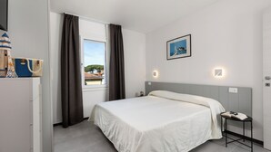 Et hotelværelse med en seng, et natbord med en bog og et par briller, en vægmonteret lampe, et billede i ramme og et vindue med gardiner.