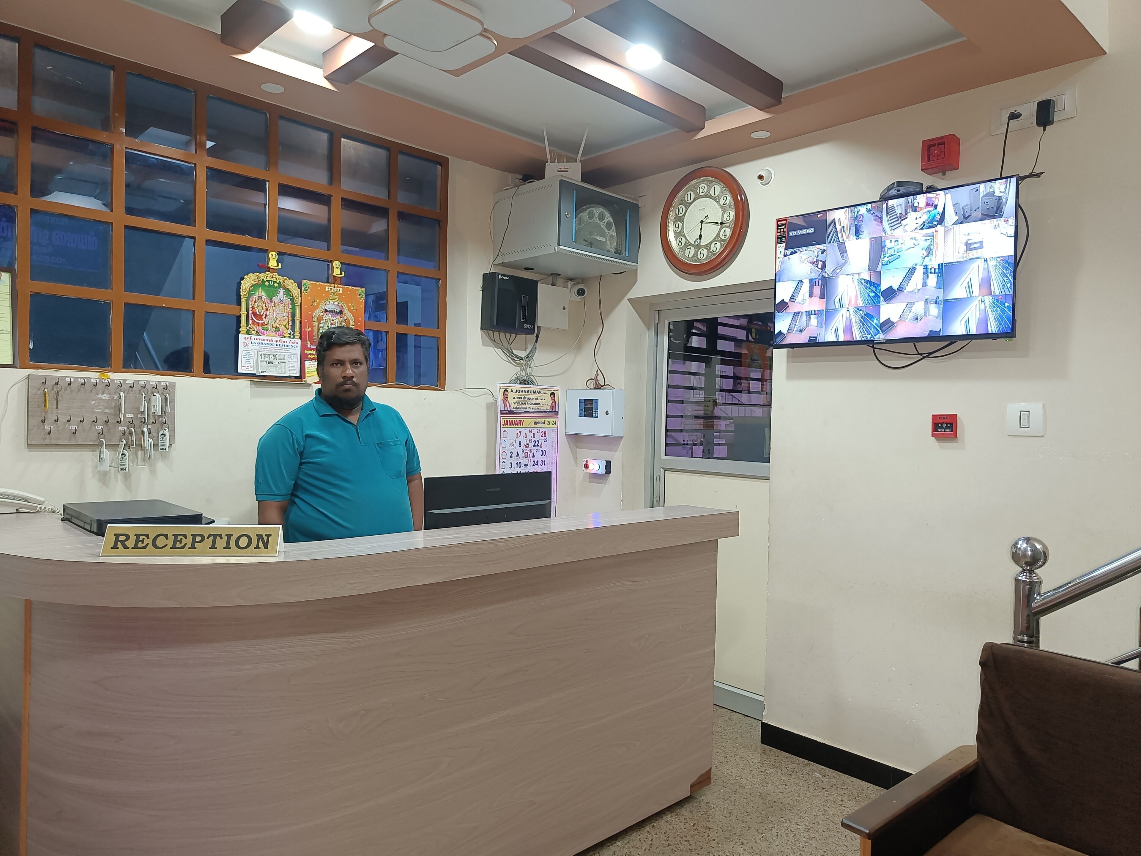 A reception desk with a clock, a wall-mounted TV displaying multiple camera feeds, and a calendar.