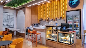 A café interior with a wooden counter, orange chairs, a display case with pastries, and a sign reading "M-NH-TT-N CAFE". - OZPED Flats Particulares Manhattan (Brasília)