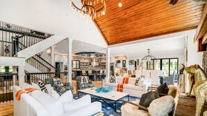 A spacious living area with a staircase, white furniture, and a kitchen in the background. - Mountain Mansion 7BR, 10Ksf, Views, Pool, Sport Court, Gym, Sauna, Petting Zoo (Blue Ridge)