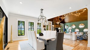 A modern dining area with a white table and chairs, a chandelier, and a view of the outdoors through large windows. - Mountain Mansion 7BR, 10Ksf, Views, Pool, Sport Court, Gym, Sauna, Petting Zoo (Blue Ridge)