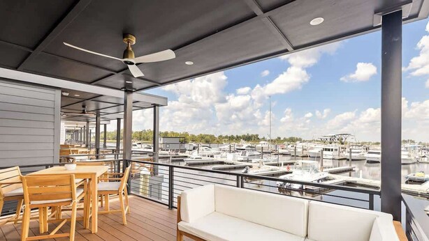 Un espacio al aire libre techado con un deck de madera, asientos y vista a un puerto deportivo con barcos.