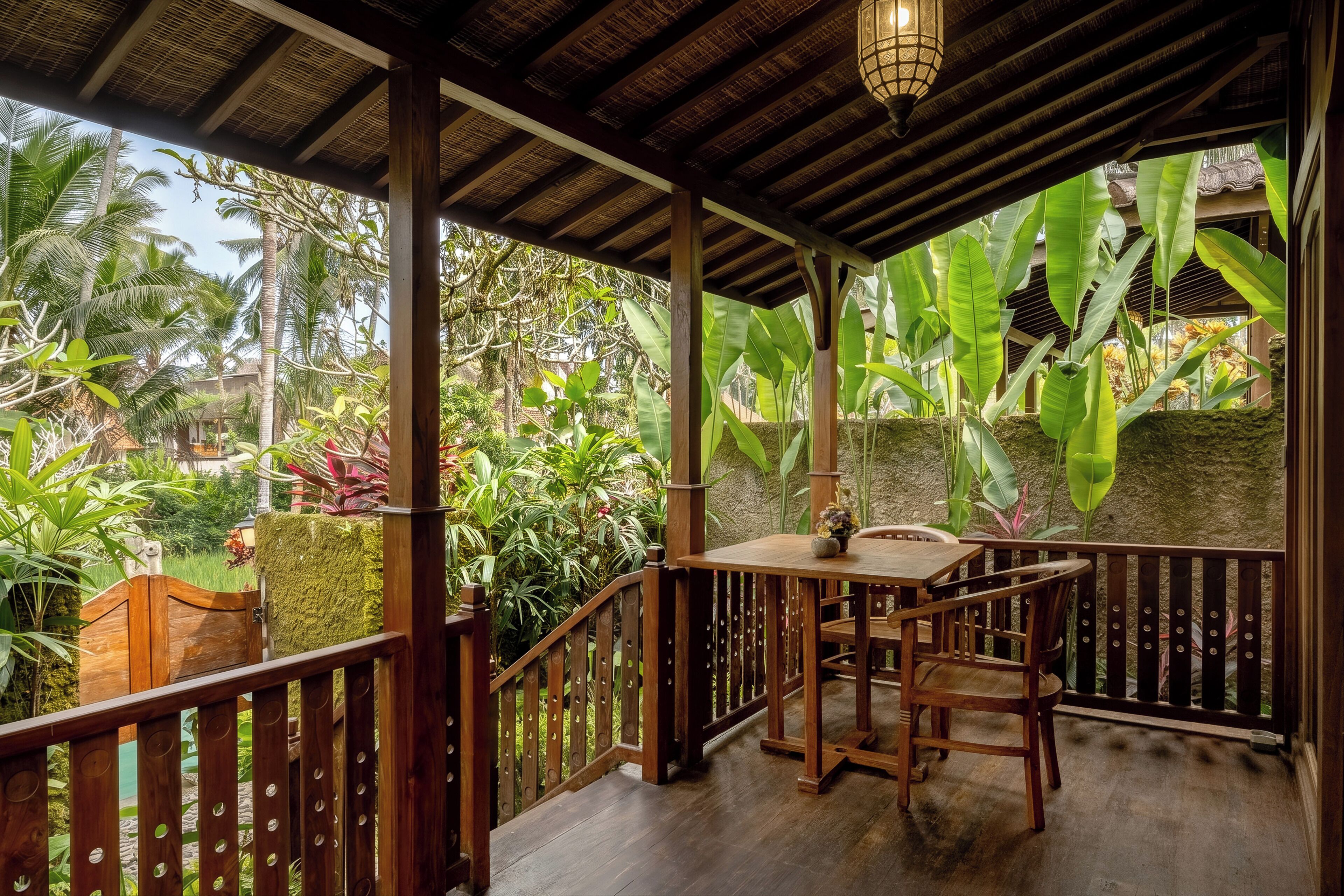 Une terrasse en bois avec une table et des chaises, entourée d’une végétation tropicale luxuriante et d’un mur de pierre.