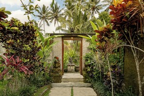 A stone pathway leading to a wooden archway surrounded by lush tropical vegetation and palm trees. - Keraman Suite Ubud (Tegallalang)