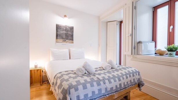 A neatly arranged bedroom with a bed, a bedside table with a lamp, a window with a view, and a wall-mounted artwork. - Taipas Tranquility: Cozy T0 by LovelyStay (Porto)