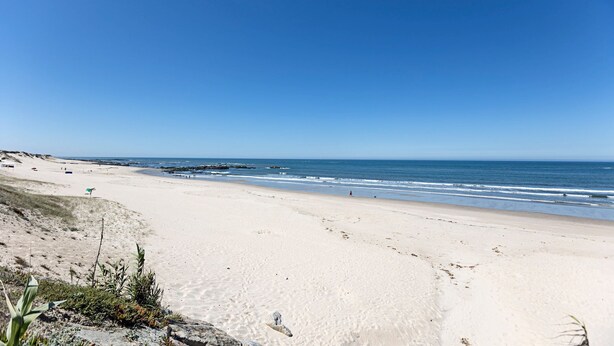 A wide sandy beach with gentle waves, a clear sky, and sparse vegetation. - Mindelo Villa Beach by LovelyStay (Vila Chã)
