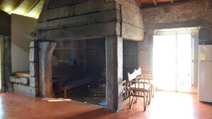 Ein rustikaler Kamin aus Stein mit einer Sitzbank, Holzstühlen und einem Kühlschrank in einem Raum mit Fliesenboden und geneigter Decke.