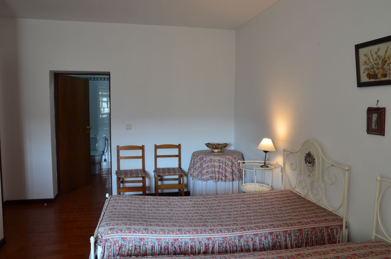 A bedroom with a bed, two chairs, a small table with a lamp, and a picture on the wall.