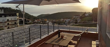 Una terraza con mesa y sillas, una sombrilla y vistas a un pueblo y colinas al fondo.