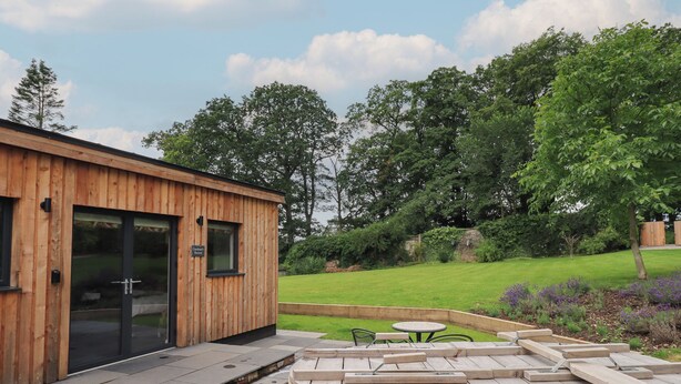 A wooden cabin with a glass door, a wooden hot tub, and a small outdoor seating area. - The Gardener's Retreat (Penrith)