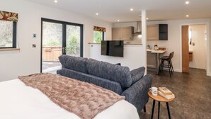 A modern living room with a sofa, a coffee table, and a TV. There is a kitchen area with a bar and a dining table. - The Gardener's Retreat (Penrith)