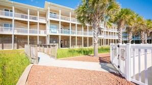 A multi-story hotel with balconies, a gravel pathway, and palm trees. - Beachside Breeze: Modern Condo in Walking Distance to Beach, Dining, Shops (Oak Island)