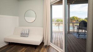 A bedroom with a bed, a round mirror, and a sliding glass door leading to a balcony with a pool and outdoor furniture. - Beachside Breeze: Modern Condo in Walking Distance to Beach, Dining, Shops (Oak Island)