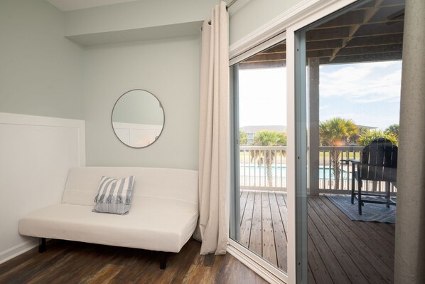 A bedroom with a bed, a round mirror, and a sliding glass door leading to a balcony with a pool and outdoor furniture.