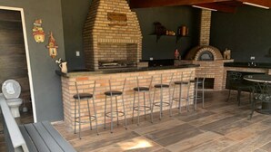 Un área de cocina al aire libre con horno de ladrillo, taburetes de bar y una mesa de comedor.