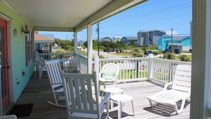 A covered porch with white rocking chairs and a table, overlooking a street with houses and parked cars. - Vitamin Sea, Almost Oceanfront House in Emerald Isle, NC! (Emerald Isle)