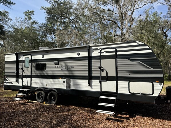 Una caravana Transcend Xplor estacionada al aire libre, con una puerta lateral, ventanas y una pequeña plataforma de acceso.