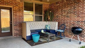 A covered patio with a brick wall, a blue sofa, a table, a chair, a grill, and a ceiling fan.