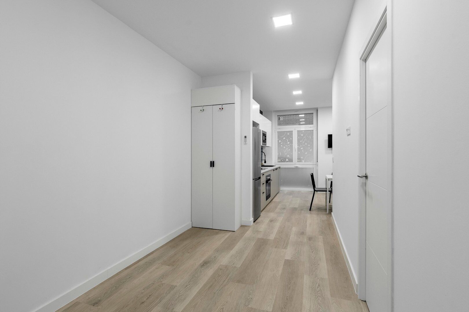 A narrow hallway with a wooden floor, white walls, and a kitchen area with a refrigerator, microwave, and stove.