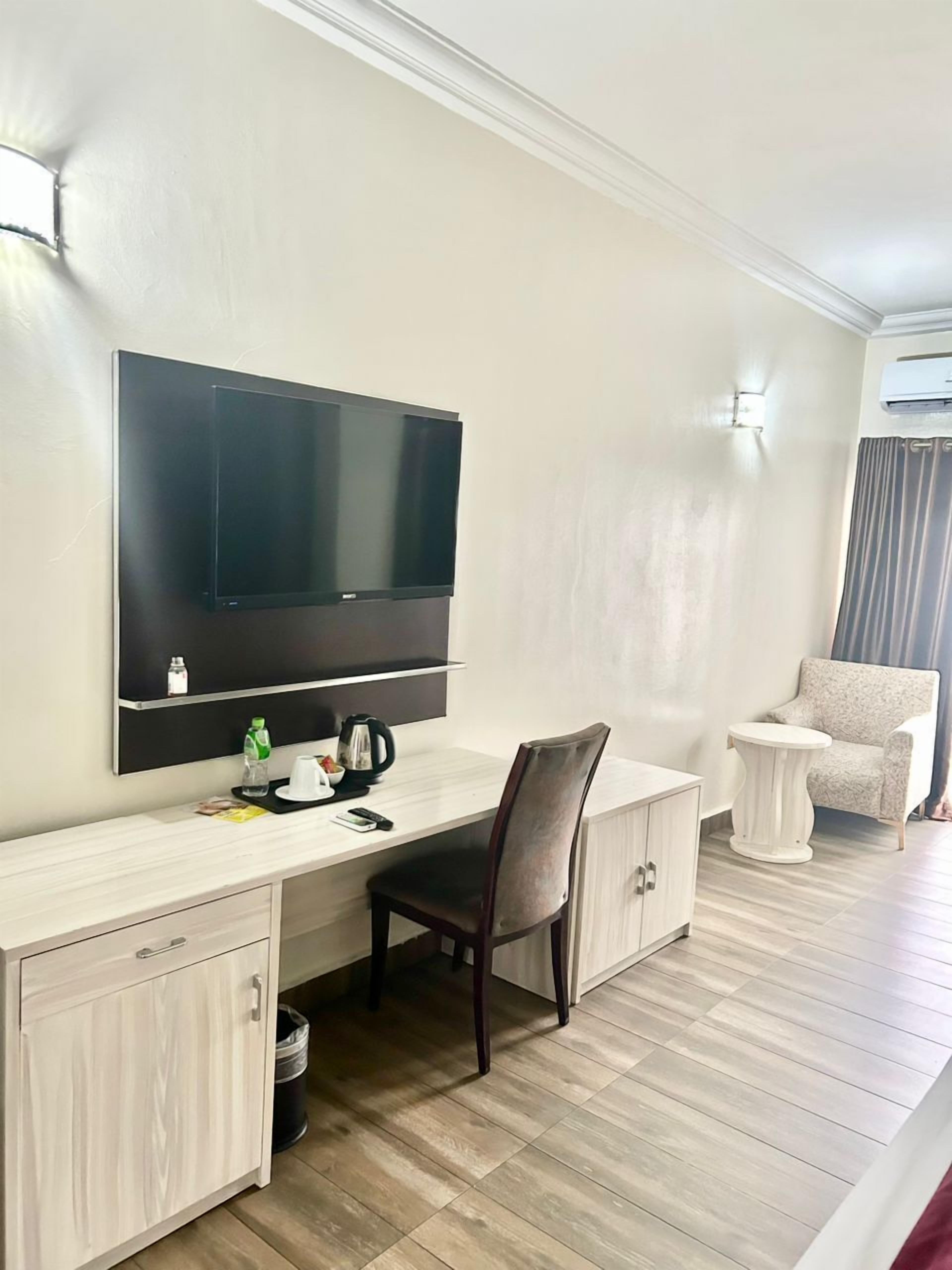 A hotel room with a large flat-screen TV mounted on the wall, a wooden desk with a chair, a small round side table, a white armchair, and a trash bin.