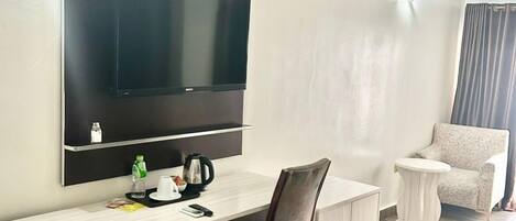 A hotel room with a large flat-screen TV mounted on the wall, a wooden desk with a chair, a small round side table, a white armchair, and a trash bin.