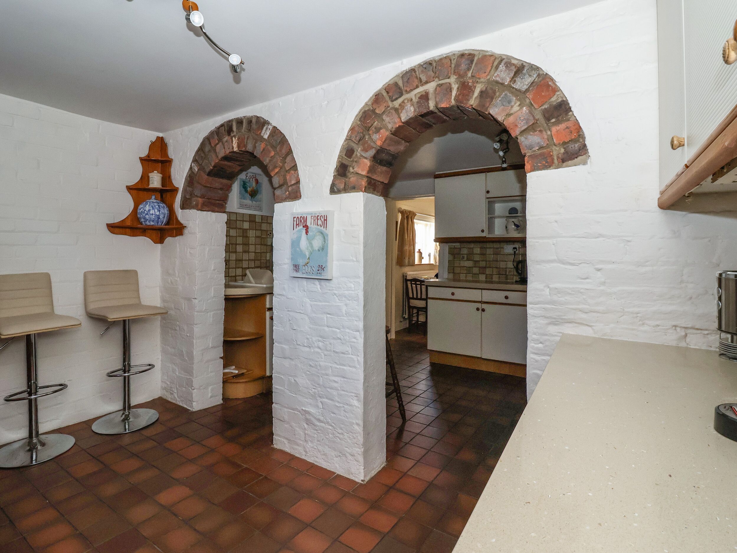 A kitchen with white brick walls, a tiled floor, and a rustic archway. There are two bar stools and a wooden shelf with a ceramic pot.