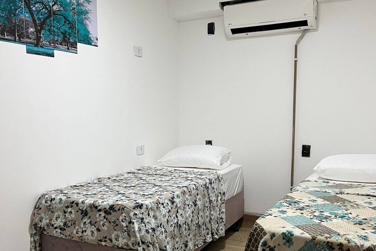 A small, clean room with two single beds, a wooden floor, white walls, and an air conditioning unit.