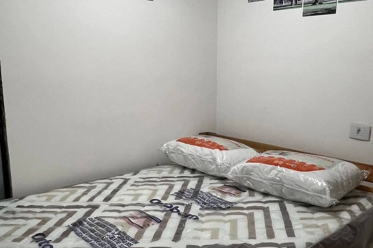 A small, single-bed room with a patterned mattress, a wooden headboard, and a wall-mounted light switch.